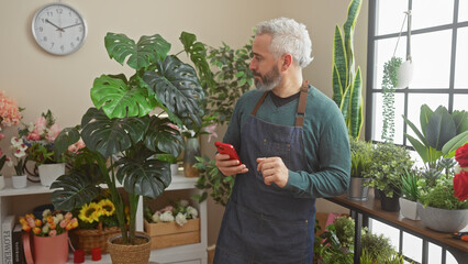 Mature man with beard in apron using smartphone amidst lush plants in a florist shop.