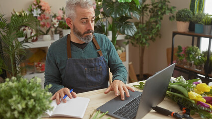 A bearded man with a laptop in a flower shop, writing notes, surrounded by plants and flowers.