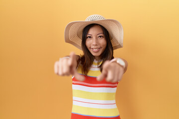 Middle age chinese woman wearing summer hat over yellow background pointing to you and the camera...