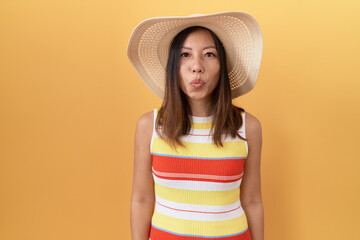 Middle age chinese woman wearing summer hat over yellow background looking at the camera blowing a...