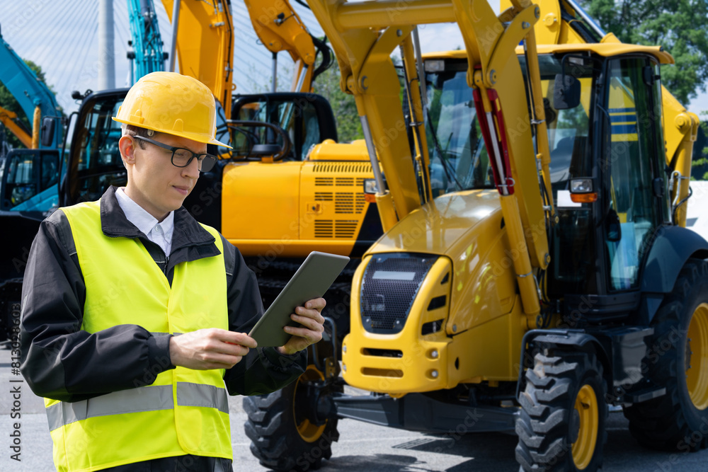 Poster Engineer in a helmet with a digital tablet stands next to construction excavators..