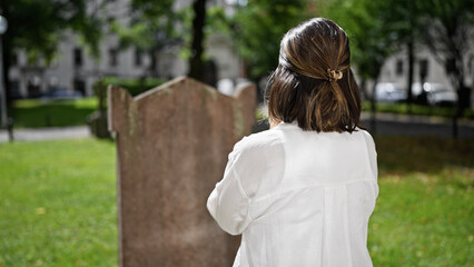 Beautiful young hispanic woman praying and mourning by tombstone at graveyard