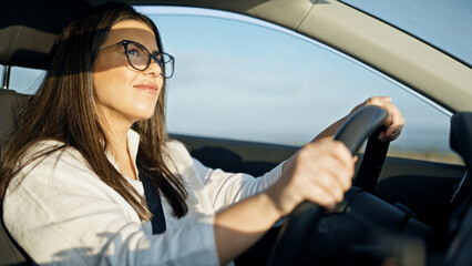 Young beautiful hispanic woman driving a car smiling wearing glasses on the road