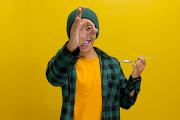 Excited young Asian man, dressed in a beanie hat and casual shirt, samples food with a spoon,...