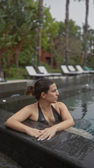 A contemplative young woman relaxes in a luxurious outdoor pool at a balinese resort, surrounded by tropical foliage.