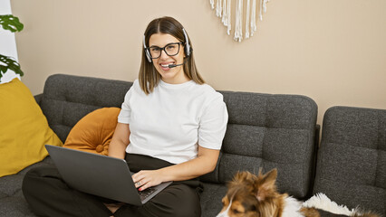 A cheerful young hispanic woman in glasses and headset uses a laptop in a cozy living room with her...