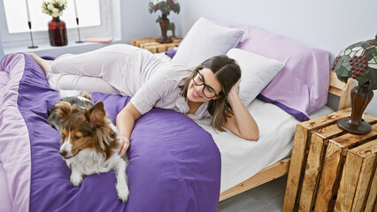 Smiling young woman with glasses resting in bed with a small dog in a cozy bedroom setting.