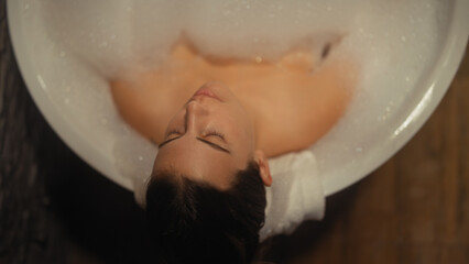 A relaxed young woman enjoys a bubbly bath indoors, her tranquility accentuated by soft lighting...