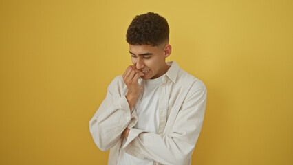 A thoughtful young man in casual attire stands against a solid yellow background, looking down with a pensive expression.
