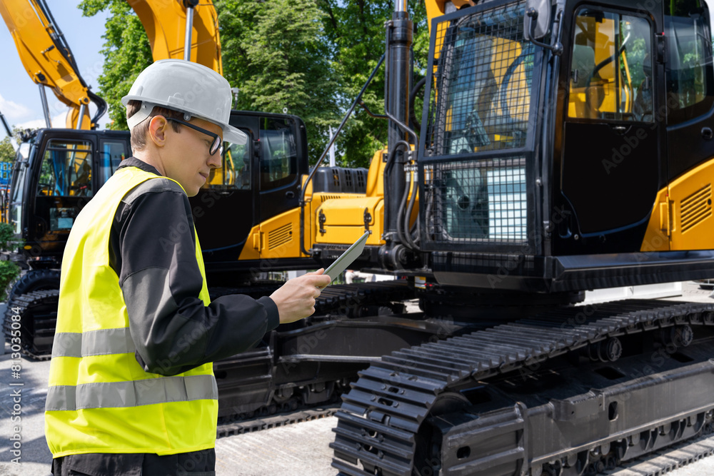 Poster Engineer in a helmet with a digital tablet stands next to construction excavators..