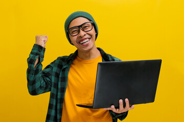 Happy young Asian man, dressed in a beanie hat and casual shirt, pumps his fist in excitement while...