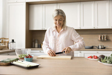 Happy senior chef woman promoting healthy nutrition, vegetarian diet, preparing salad for dinner, chopping fresh vegetables at table with natural ingredients, enjoying culinary hobby, smiling