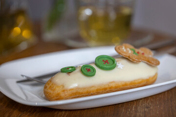  festive Christmas-themed Ã©clair adorned with a gingerbread man and green buttons, set on a white plate with a silver fork. The blurred background suggests a dining table with a glass of water