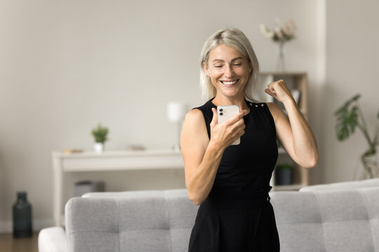 Cheerful mature blonde pretty woman reading exciting message on smartphone, looking at screen, making winner hand gesture, celebrating success, achievement, luck, dancing, laughing
