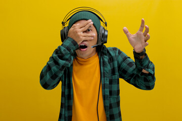 Asian man in a beanie and casual clothes, wearing headphones, peeks through his fingers in shock after hearing something embarrassing on his music. Isolated on a yellow background.