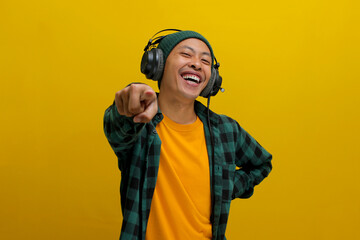 Excited Asian man in a beanie and casual clothes, wearing headphones, points excitedly towards the...