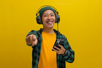 Asian man in a beanie and casual clothes points his finger directly at the camera while listening to music on his headphones. His phone is in his hand. Isolated on a yellow background.