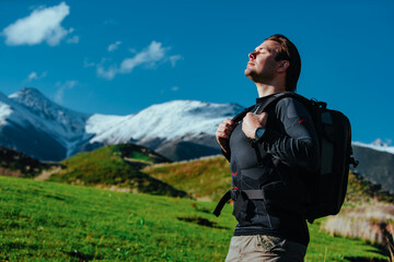 Man hiker with backpack stands in picturesque valley on mountains background with closed eyes