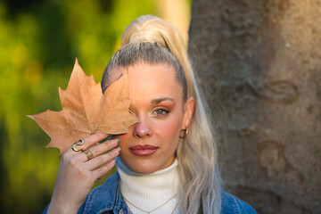 Pretty young blonde woman with a ponytail in her hair covers one eye with a dry leaf. Focus on the...