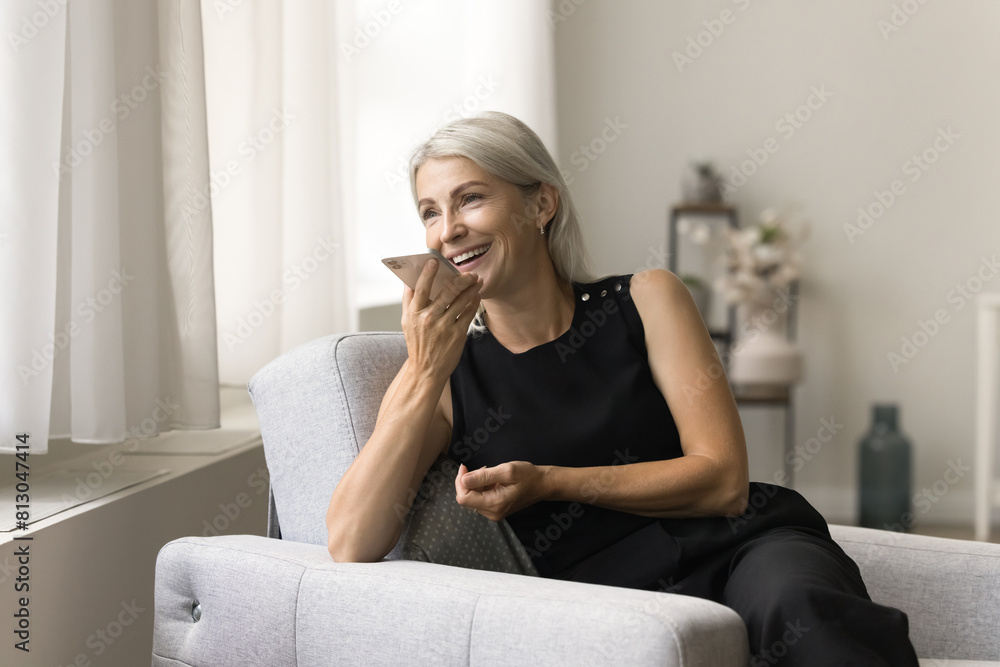 Wall mural cheerful blonde senior woman enjoying leisure, mobile phone communication in cozy armchair, talking 