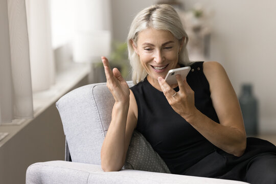 Positive mature lady talking on speaker on mobile phone call, holding digital gadget at face, speaking with hand gestures, recording voice message, sitting in armchair, enjoying communication