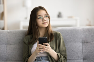 Serious pensive beautiful teenager girl in stylish glasses holding cellphone, looking away with...