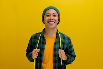 Cheerful Asian man in a beanie and casual shirt smiles at the camera, holding a yellow measuring tape around his neck. Isolated on a yellow background