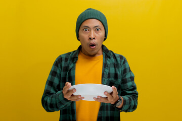 Surprised Asian man, dressed in beanie hat and casual shirt, holds an empty white plate in his...
