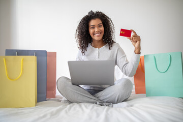 A Hispanic woman shopaholic is seated on a bed, holding a credit card in one hand and a laptop in...