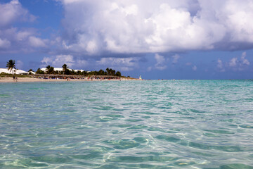 Defocused view from ocean surface to tropical beach with people, sandy coast and coconut palm trees. Sea resort on Caribbean island with clear water