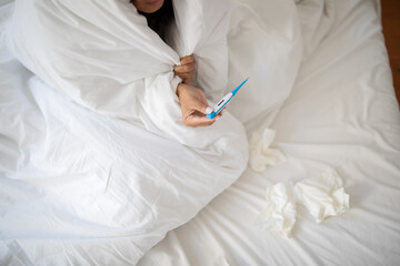 Cropped of woman appears to be sick, tucked under a white blanket, holding a thermometer to check...