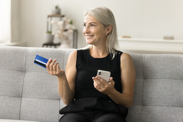 Positive satisfied senior bank customer woman sitting on home couch with blue credit card and...