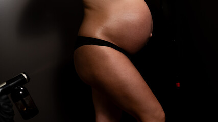 Faceless pregnant woman undergoing an instant tanning procedure. The master applies bronzer to the legs with a spray. 