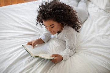 Hispanic woman is lying comfortably on a bed, engrossed in a book she is reading. She appears...