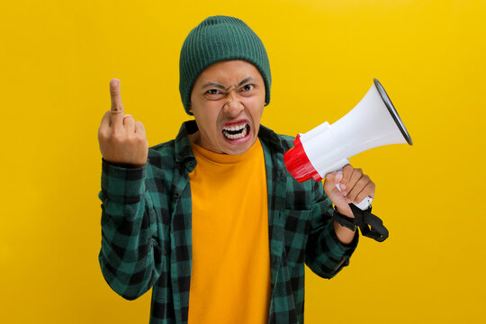 Angry Asian man in a beanie hat shouts and yells into a megaphone while showing his middle finger at the camera, demanding change, attention, or protesting. Isolated on a yellow background