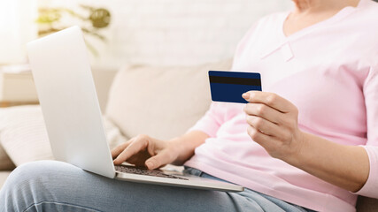 Cropped of woman is seated on a couch, holding a credit card in one hand and a laptop in the other....