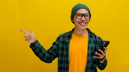 Asian man wearing beanie hat, casual shirt, and eyeglasses points at an empty space while...
