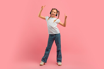 A young girl wearing headphones stands in front of a vibrant pink background. She appears engaged in listening to music and dancing