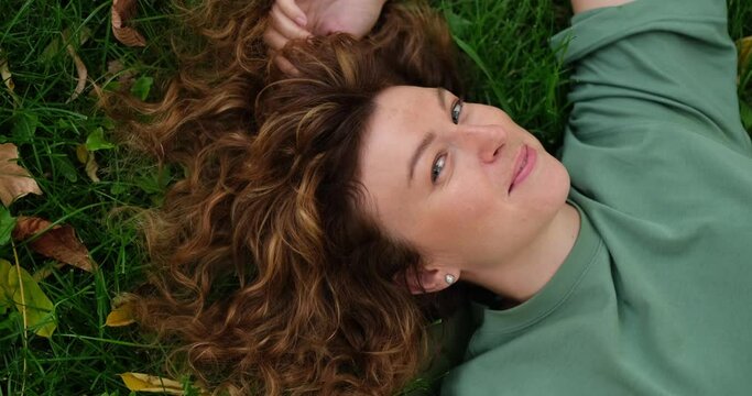 Redhead girl lying on green grass with positive emotions, smiling and feeling happy. Beautiful woman at sunny summer luing in park outdoors. Happy positive concept, happiness emotions