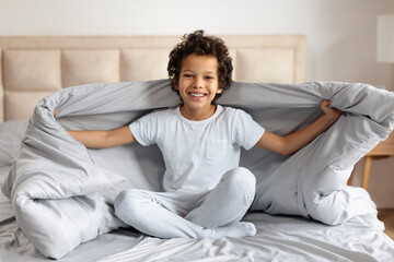 Fototapeta premium A cheerful young African American boy sits cross-legged on a bed, spreading a soft blue blanket with both hands in a warmly lit bedroom.