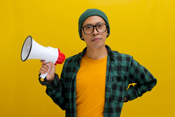Confident Asian man in a beanie hat and casual shirt holding a megaphone, isolated on a yellow...