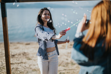 A cheerful young woman blows bubbles towards her friend who is capturing the moment on camera,...