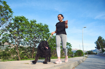 Happy woman walking her dog on leash in the nature. Smiling female in sportswear, enjoying walk...