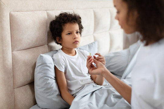 African American young child sits with a contemplative expression on bed while an adult, partially visible, holds out a pill for him to take, likely indicating a moment of healthcare at home. - Powered by Adobe