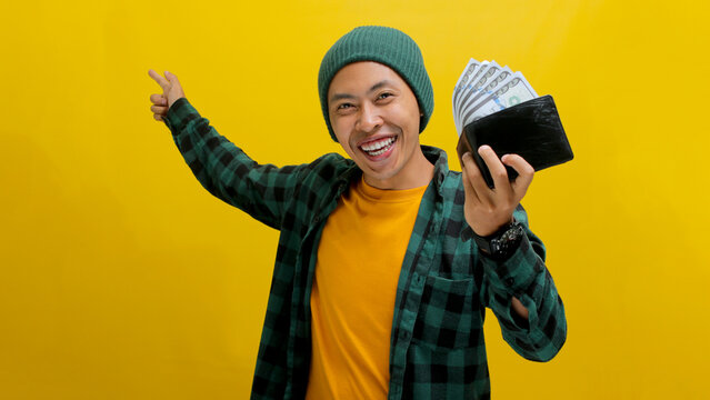 An enthusiastic young Asian man, dressed in a beanie hat and casual shirt, points to an empty space for text or advertisement while holding a wallet, isolated on a yellow background