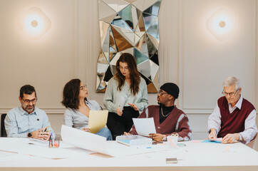 A multi-ethnic team of professionals collaborates around a table full of documents and laptops,...