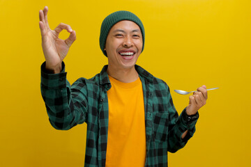 Excited young Asian man, dressed in a beanie hat and casual shirt, samples food with a spoon,...