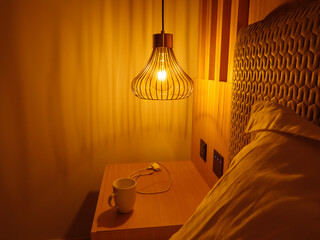 Metal lamp shade with yellow light above bedside cabinet with the cup and type-c charging device on the background and blurry pillow on the foreground. Relaxing evening atmosphere concept.