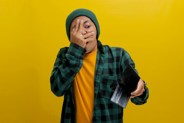 Funny young Asian man sporting a beanie hat and casual shirt covers his face and peeks through his fingers while observing his empty wallet, humorously depicting financial difficulties and being broke