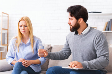Bearded man psychologist offer glass of water to upset distressed young blonde woman client, both...